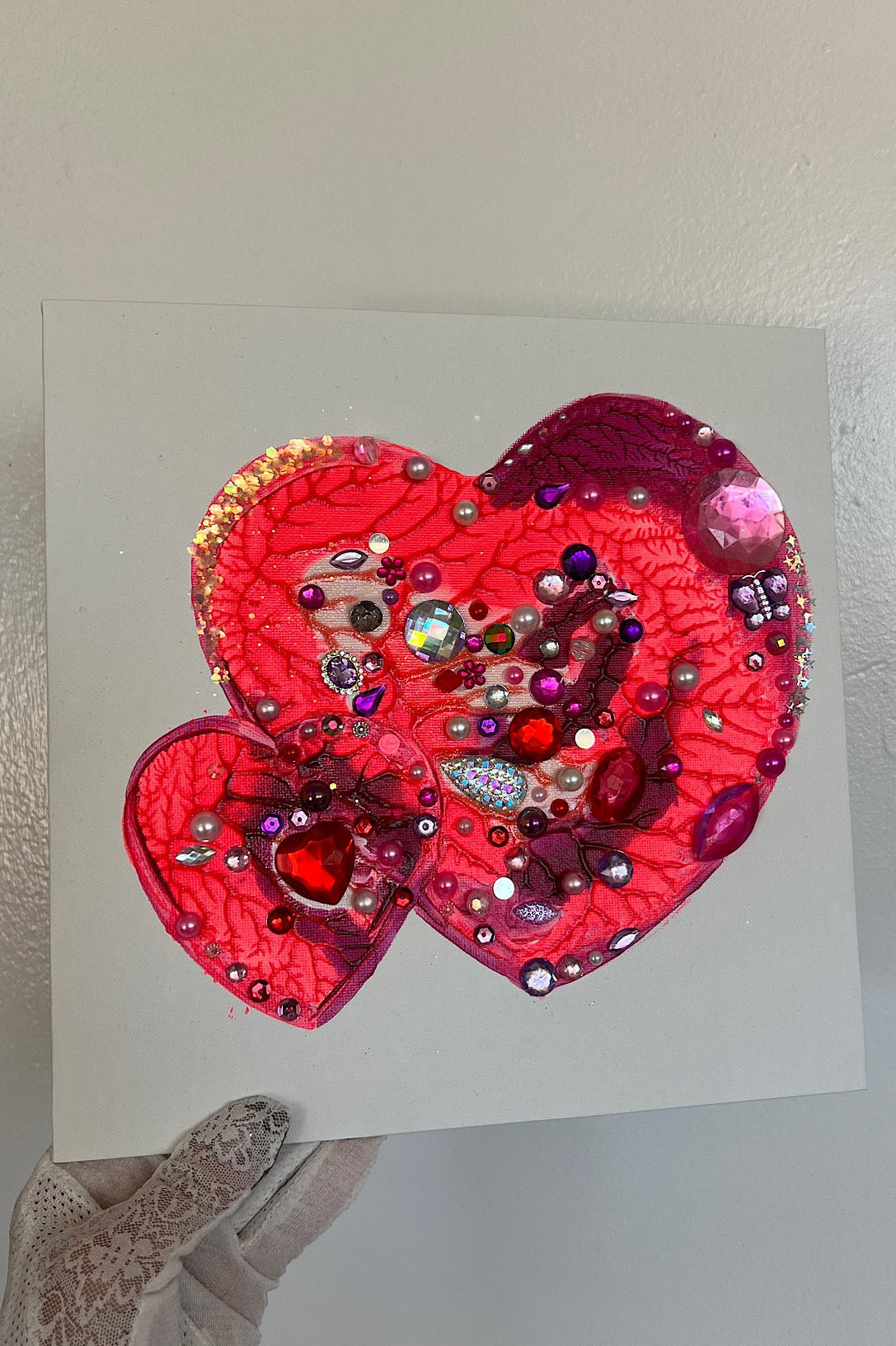 Decorative heart-shaped object with sequins on a white card, held by a gloved hand.