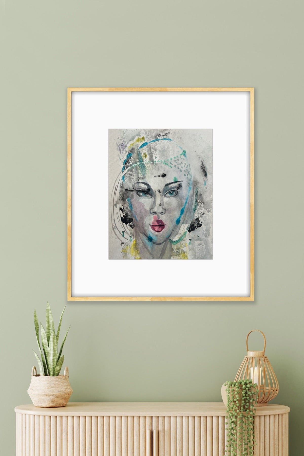 Framed mixed-media portrait artwork hanging on a sage green wall, styled with a snake plant and lantern decor on a light wood credenza