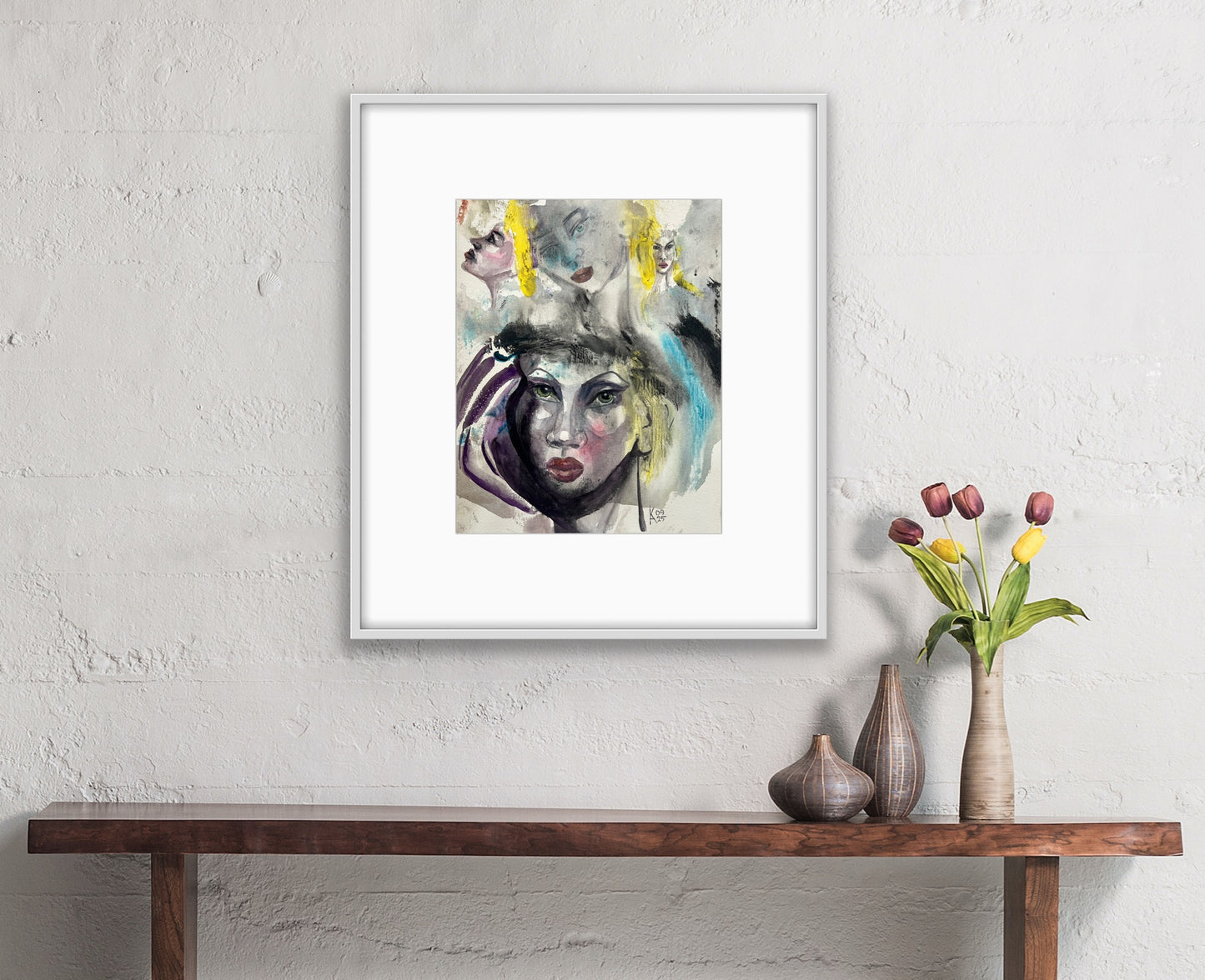Framed abstract watercolor portrait displayed on a white brick wall above a wooden console table with vases and tulips.