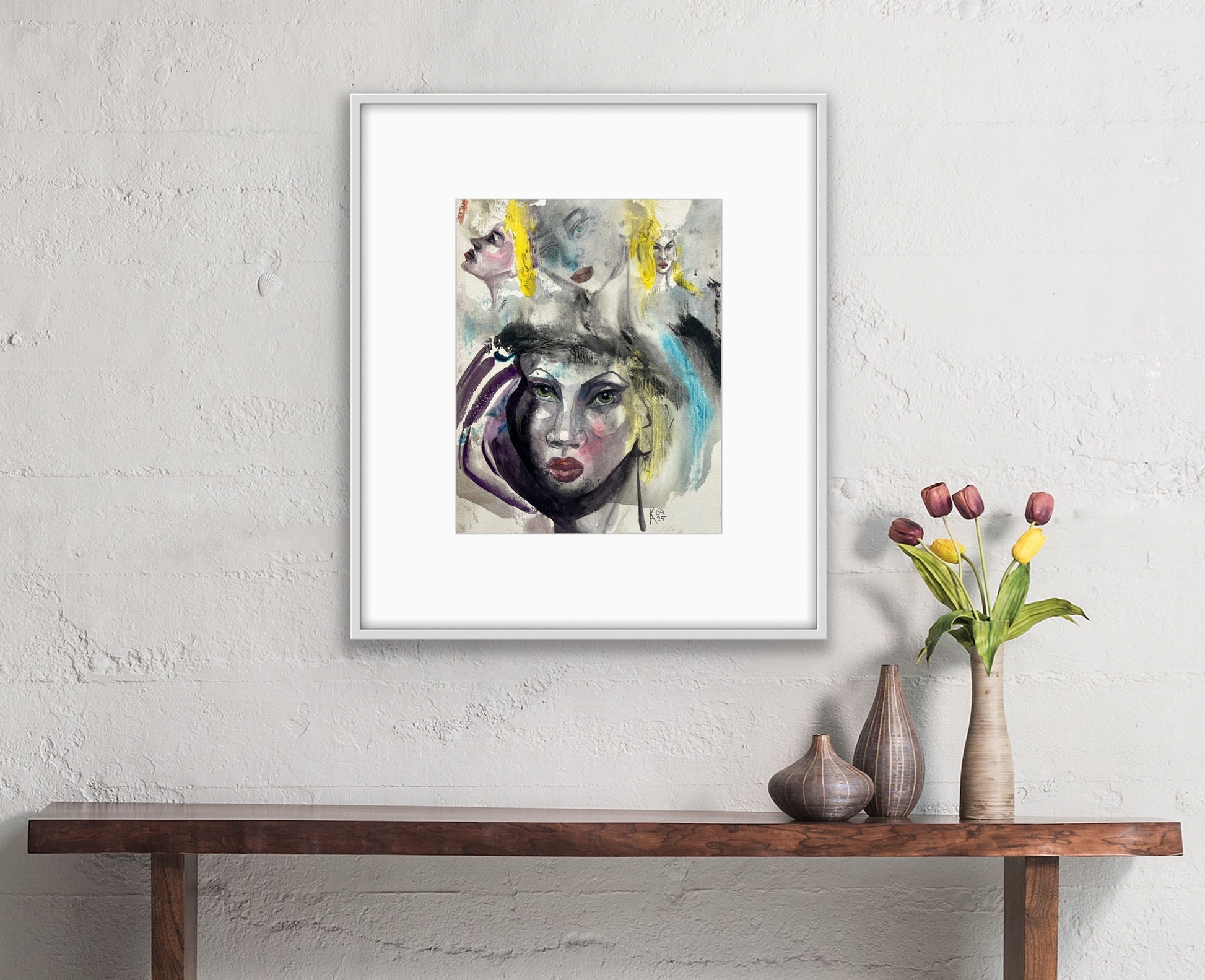 Framed abstract watercolor portrait displayed on a white brick wall above a wooden console table with vases and tulips.