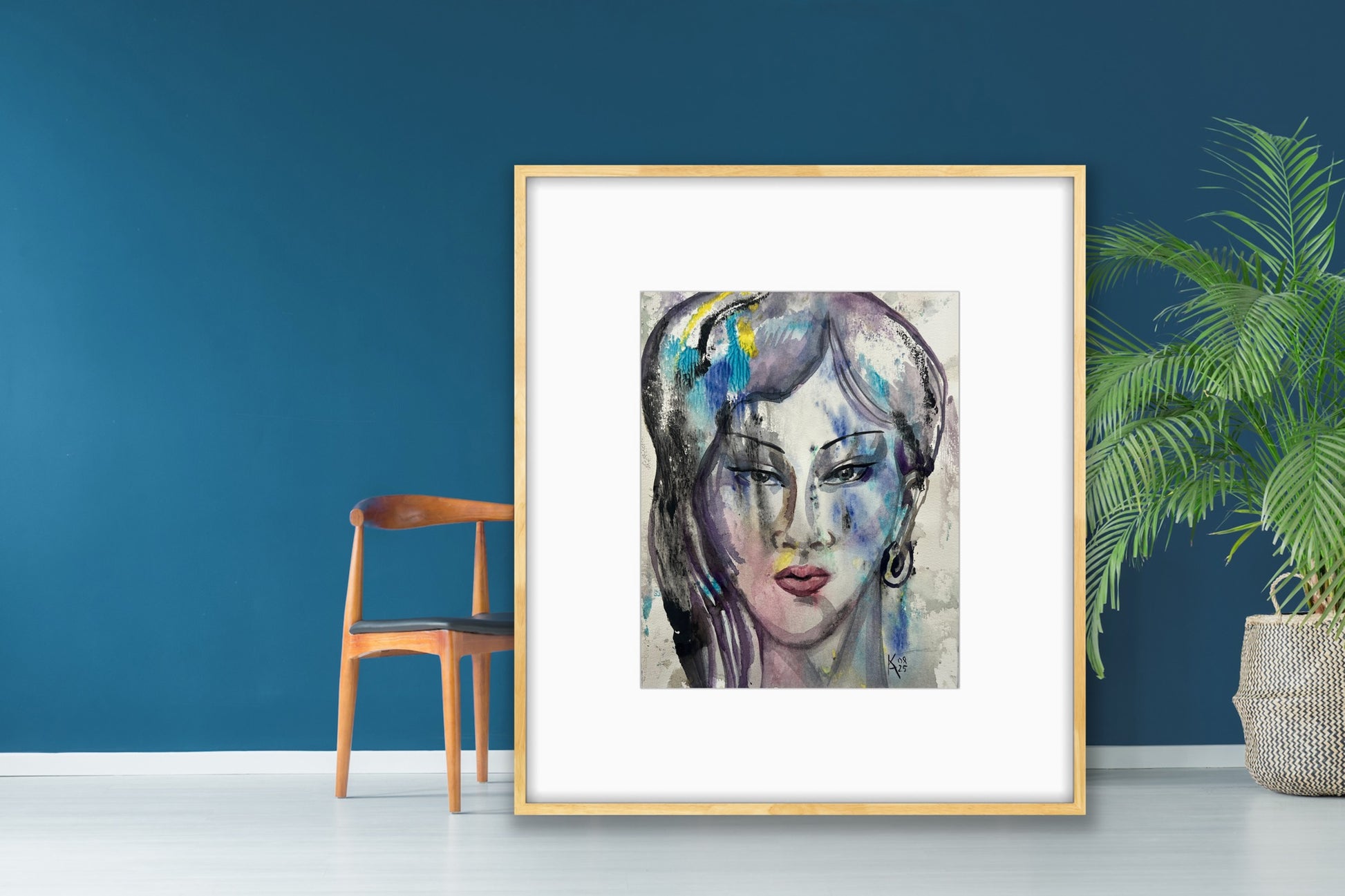 Large light wood framed watercolor abstract portrait leaning casually against a dark blue wall on a white floor, paired with a wooden chair and a potted green plant, creating a relaxed modern art vignette