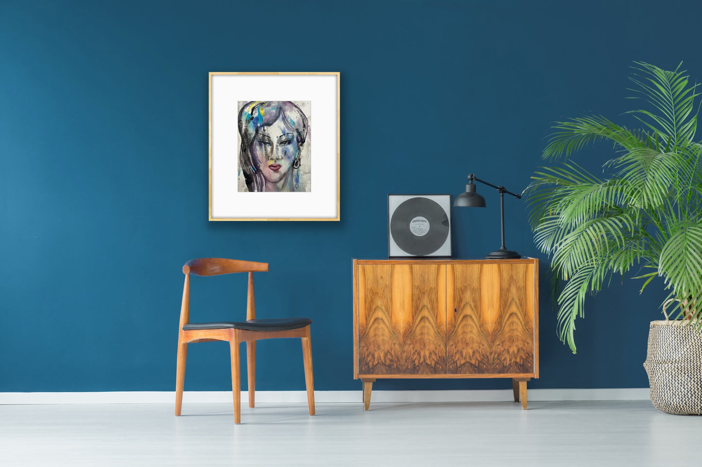 Framed watercolor portrait artwork displayed on a deep teal blue wall above a wooden mid-century cabinet, styled with a vinyl record player, black lamp, and a potted palm plant for an elegant retro decor setup